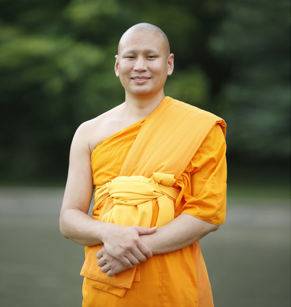 Luang Phi Pasura vistiendo una túnica naranja con un bosque detrás.